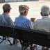 Three elderly men sitting on a park bench, viewed from behind, wearing hats and light-colored shirts. - Olive Oil Times