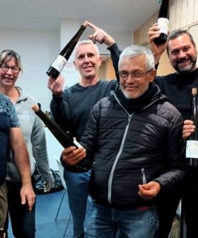Six individuals posing together while holding bottles of olive oil in a workshop setting. - Olive Oil Times