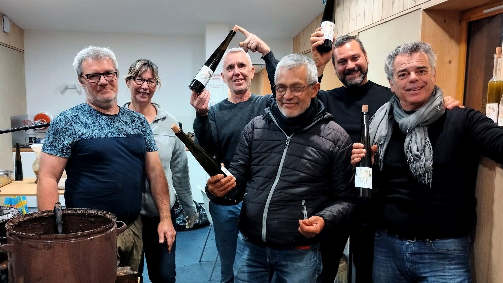 Six individuals posing together while holding bottles of olive oil in a workshop setting. - Olive Oil Times