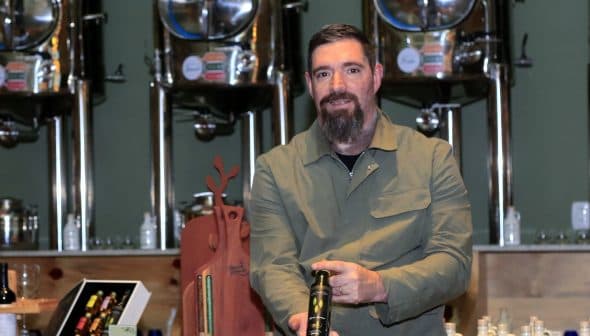 Man with a beard holding a bottle of olive oil in a production facility with metal containers in the background. - Olive Oil Times