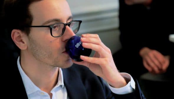 Man wearing glasses drinking from a blue glass with the word 'Olive' on it. - Olive Oil Times
