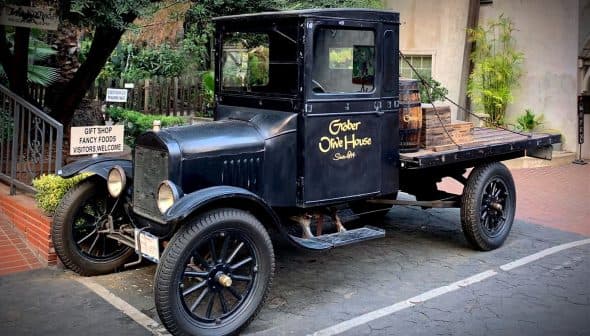 A vintage black pickup truck with wooden barrels in the back, parked on a paved surface. - Olive Oil Times
