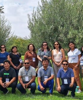 A group of 14 people standing on grass with trees in the background. Some are wearing name tags and sunglasses. The setting appears to be a park or garden.