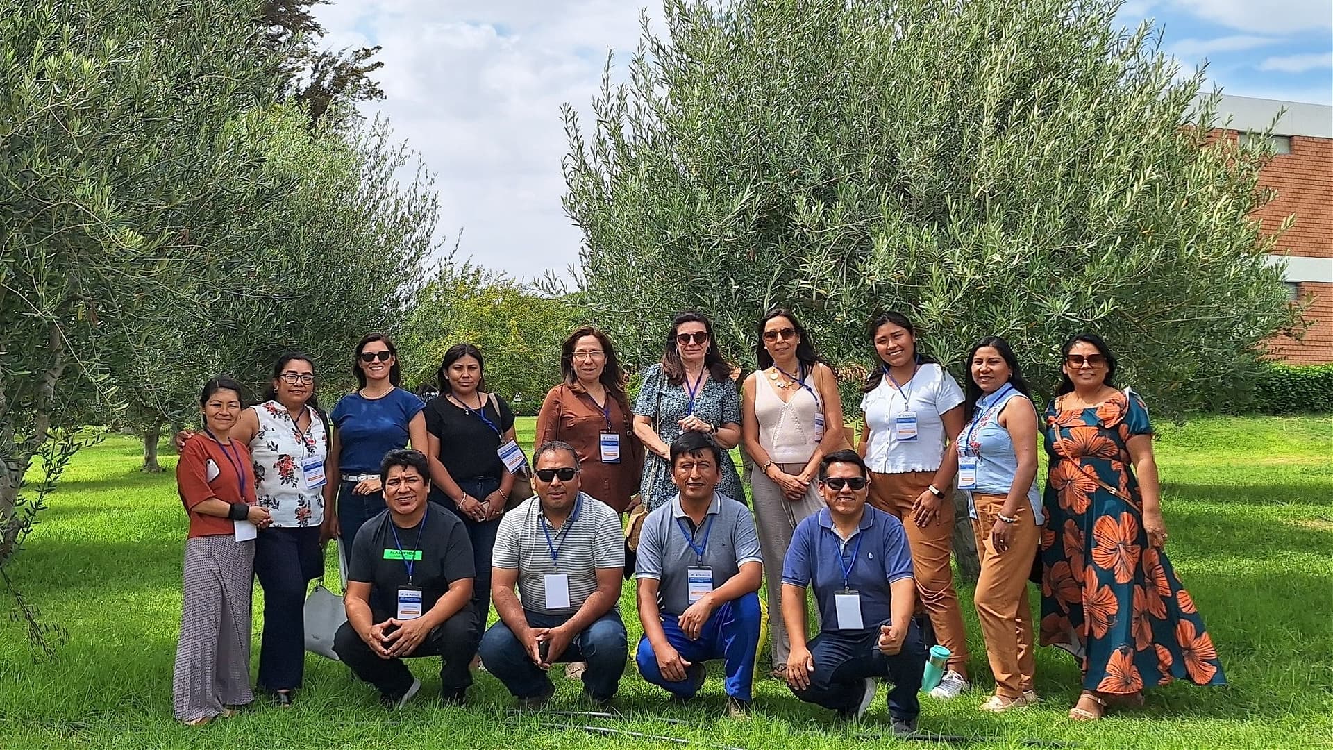 A group of 14 people standing on grass with trees in the background. Some are wearing name tags and sunglasses. The setting appears to be a park or garden.