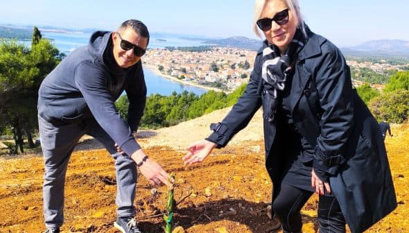 Two individuals bending down to plant a small tree in a rural area with a view of distant hills and water. - Olive Oil Times
