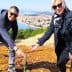 Two individuals bending down to plant a small tree in a rural area with a view of distant hills and water. - Olive Oil Times