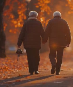 An elderly couple, dressed in warm coats, walks hand in hand down a path covered with autumn leaves. The sun sets in the background, casting a warm, golden light over the scene. The woman holds a small handbag.