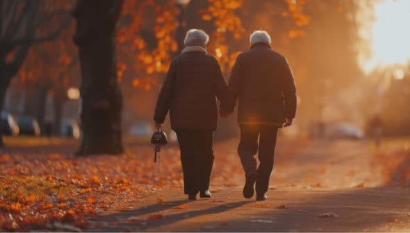 An elderly couple, dressed in warm coats, walks hand in hand down a path covered with autumn leaves. The sun sets in the background, casting a warm, golden light over the scene. The woman holds a small handbag.