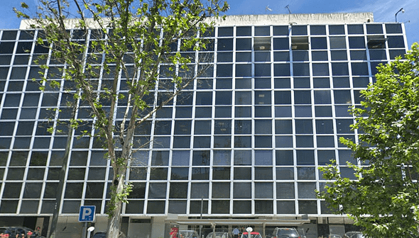 Ένα μοντέρνο κτίριο γραφείων με πλέγμα από μεγάλα παράθυρα και ένα δέντρο μπροστά. - Olive Oil Times