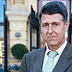 Un homme portant un costume marron et une cravate verte, debout à l'extérieur près de l'entrée d'un bâtiment. - Olive Oil Times