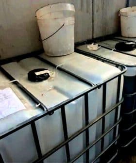 Two large industrial storage tanks with a white bucket placed on top in a warehouse setting. - Olive Oil Times