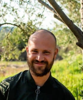 Man with a beard smiling while standing between olive trees in a natural setting. - Olive Oil Times