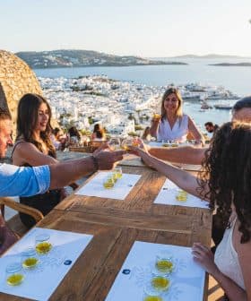 A group of five people toasting with drinks at a wooden table overlooking a coastal view. - Olive Oil Times